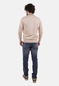 Maglione beige chiaro da uomo con collo rotondo, polsini e orlo a costine, abbinato a jeans blu e scarpe da ginnastica marroni. Dettagli visibili sul retro.