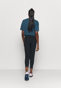 Geknipte teal t-shirt en zwarte strakke broek, gecombineerd met donkerblauwe sportschoenen met roze accenten, gezien van achteren.