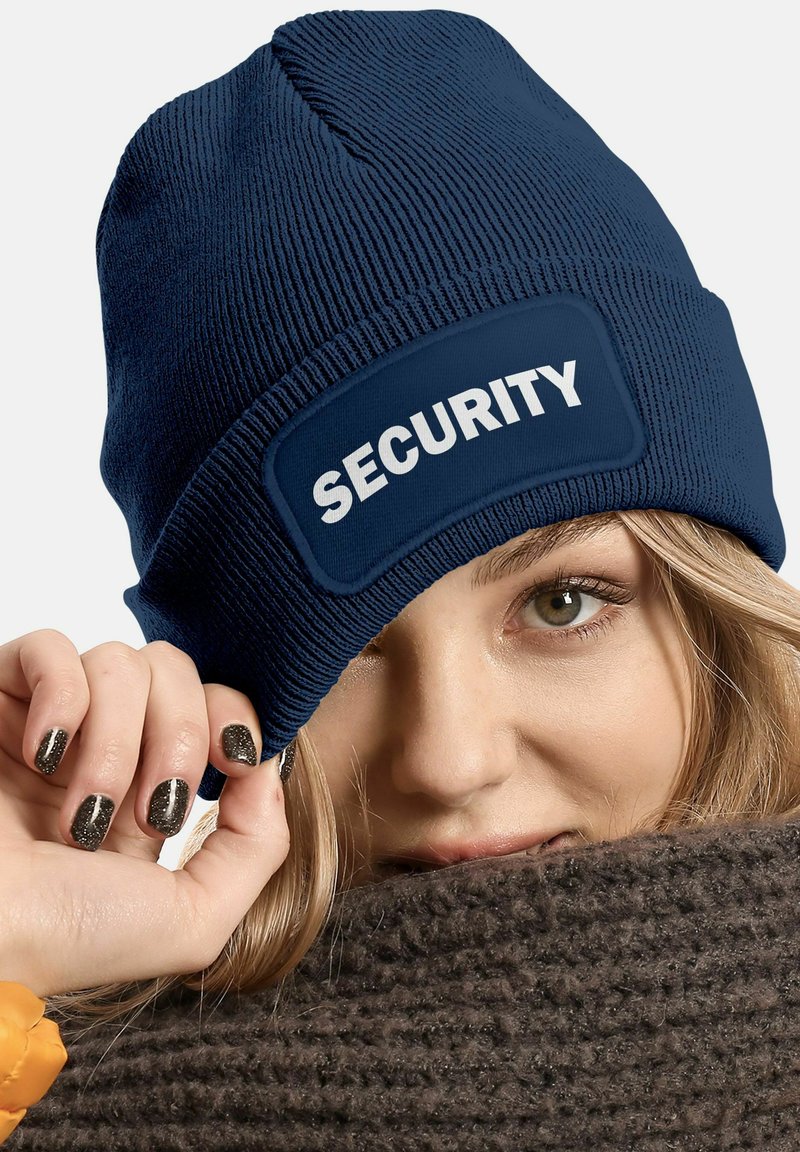 Navy beanie made of ribbed fabric featuring a white "SECURITY" text patch. Soft texture; snug fit. Hand on textured brown scarf.