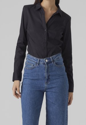 Camicia nera con bottoni, realizzata in tessuto liscio, caratterizzata da maniche lunghe e colletto strutturato, abbinata a jeans in denim blu a vita alta.