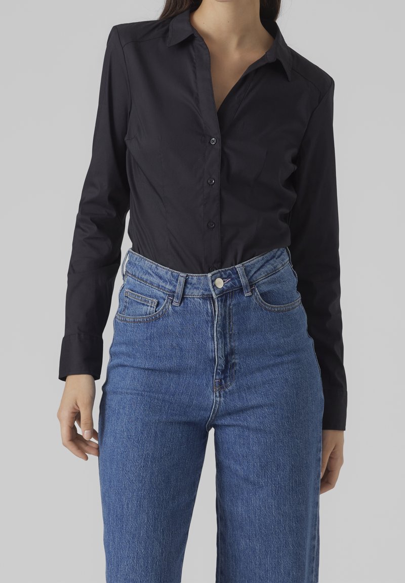 Camicia nera con bottoni, realizzata in tessuto liscio, caratterizzata da maniche lunghe e colletto strutturato, abbinata a jeans in denim blu a vita alta.