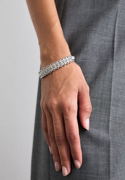 Srebrna narukvica s prozirnim rhinestones, s složenim rešetkastim dizajnom, nosi se na ruci na sivoj tkanini.