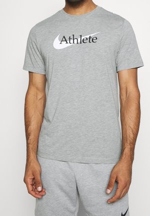 Homme portant un T-shirt gris clair à manches courtes avec le texte noir « Athlete » et le logo swoosh blanc, assorti à un short gris.