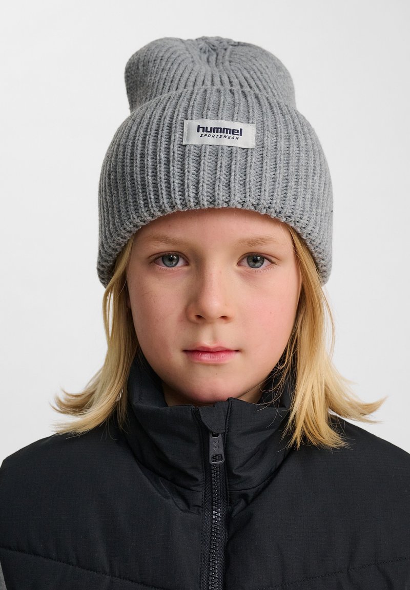 Grå stickad mössa med ribbad textur och vikbar brätte, med en vit etikett som säger "hummel sportswear." Bärs av ett barn i en svart jacka.
