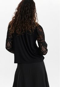 Top nero con corpetto strutturato e maniche lunghe in pizzo, caratterizzato da intricati motivi floreali e una vestibilità rilassata. Abbinato a una gonna nera.