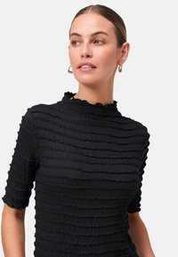 Donna con pelle chiara e capelli castani acconciati all'indietro, indossa orecchini a cerchio e una maglietta nera a maniche corte con texture, mentre guarda di lato.