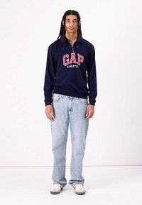 Sudadera con cremallera en color navy con el logo rojo "GAP ATHLETIC", fabricada en un material ligero, combinada con jeans rectos de color azul claro y zapatillas.