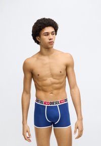 Zilas bokseršorti ar melnu un sarkanu elastīgu jostu, uz kuras ir Diesel logo. Mīksts kokvilnas materiāls, pieguļošs dizains un balti akcenti.