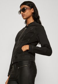 Svart denimjakke med glidelås foran, tettsittende design og sidelommer. Kombinert med svarte imitert skinnbukser. Klare solbriller fullfører antrekket.