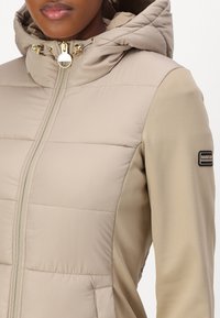 Svetle béžová péřová bunda s přiléhavým střihem, nabízí zapínání na zip, kapuci a zlaté kovové detaily. Obsahuje nášivku s logem na rukávu.