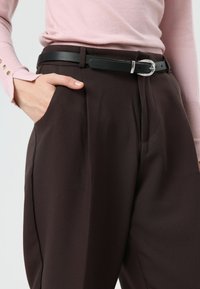 Zwarte leren riem met zilveren gesp gedragen over donkerbruine broek met hoge taille, gecombineerd met een lichtroze top met lange mouwen en knoopdetails.