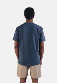 Tmavě modré bavlněné tričko s krátkými rukávy a kulatým výstřihem, nošené s béžovými šortkami, pohled zezadu. Hladká textura, volný střih.