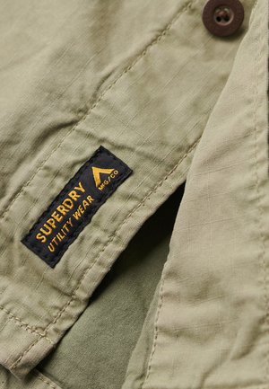 Camicia utilitaria verde con tessuto testurizzato, chiusura a bottone e un'etichetta nera con testo giallo che recita "SUPERDRY UTILITY WEAR".