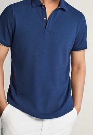 Homme portant un polo bleu marine à manches courtes avec col boutonné et un pantalon blanc, les mains dans les poches du pantalon.