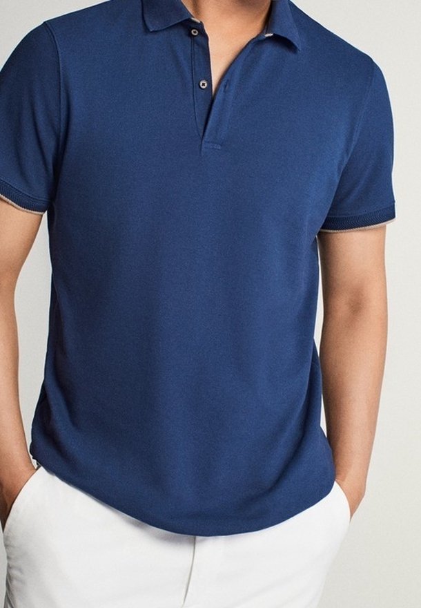 Homme portant un polo bleu marine à manches courtes avec col boutonné et un pantalon blanc, les mains dans les poches du pantalon.