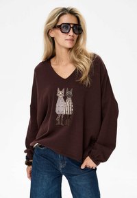 Brązowa bluza z dekoltem w serek i długimi rękawami, ozdobiona haftowanym wzorem dwóch kotów o różnych wzorach. W zestawie z niebieskimi dżinsami.