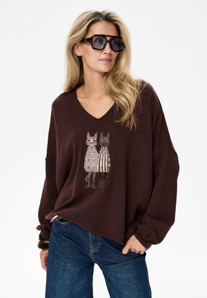 Brązowa bluza z dekoltem w serek i długimi rękawami, ozdobiona haftowanym wzorem dwóch kotów o różnych wzorach. W zestawie z niebieskimi dżinsami.