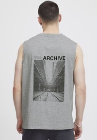 Pilnas rankovės pilkos medvilnės marškinėliai su dideliu juodu ir baltu grafikos spaudiniu, kuriame parašyta "ARCHIVE" ir tekstas virš jo.