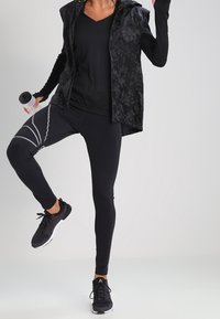 Czarny strój sportowy składający się z bezrękawnika z kapturem o teksturowanym wzorze, dopasowanej czarnej bluzki oraz czarnych legginsów z szarymi akcentami.