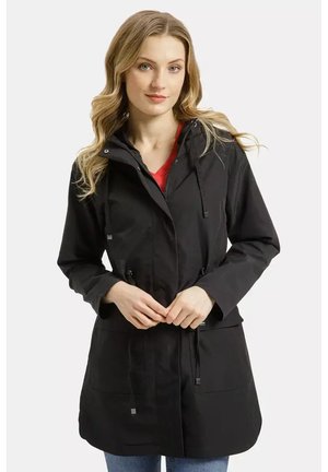 Młoda kobieta z falującymi blond włosami, ubrana w czarną kurtkę z kapturem na czerwonej bluzce, dłonie poprawiające wiązania z przodu, stoi na białym tle.