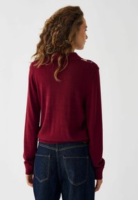 Suéter burdeos de manga larga con cuello, que presenta acentos en los hombros. Combinado con jeans de mezclilla oscuros que muestran costuras en contraste.