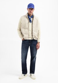 Beige fleece zip vest over een lichte trui, gecombineerd met donkere jeans en witte sneakers. Een blauwe pet en een patroon-sjaal maken de look compleet.