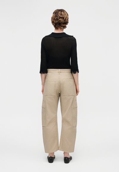 Άτομο με κοντά σγουρά μαλλιά που φοράει μαύρο μακρυμάνικο μπλουζάκι και μπεζ φαρδιά παντελόνια cargo, στέκεται με την πλάτη προς τα εμάς σε απλό φόντο.