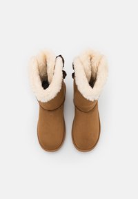 Bottines en daim marron avec doublure en fausse fourrure et bord replié. Présente un nœud décoratif de chaque côté et des semelles plates.