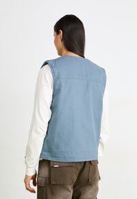 Gilet sans manches bleu en coton, à la coupe décontractée, avec des détails de couture subtils, superposé à une chemise à manches longues blanche.