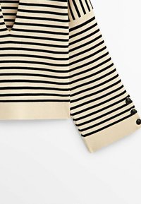 Haut en maille beige à manches longues avec des rayures horizontales noires et trois boutons noirs sur le poignet, présenté sur un fond blanc.