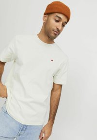 Ljusbeige bomullströja med korta ärmar och rund halsringning, med en liten röd pricken-logotyp på bröstet. Bäddad med ljusblå jeans.