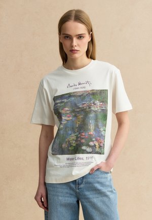 Jonge vrouw met steil blond haar, een wit T-shirt met Monet's schilderij Waterlelies uit 1916 en een blauwe spijkerbroek.