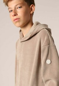 Haut à capuche beige côtelé avec un cordon de serrage, en tissu texturé, et un badge circulaire sur la manche gauche. Il présente une coupe décontractée et une poche kangourou.