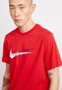 Red Nike t-shirt with a crew neckline, featuring a white swoosh logo. Material appears smooth and lightweight. Tattoo visible on the arm.