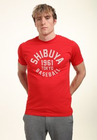Červené bavlněné tričko s bílou kruhovou grafikou a textem "SHIBUYA 1961 TOKYO BASEBALL". Krátké rukávy a kulatý výstřih.