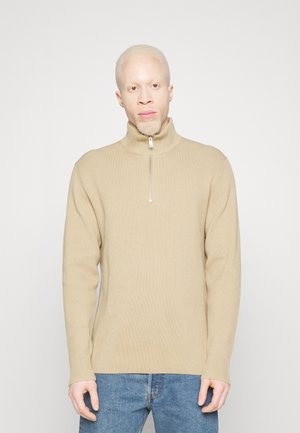 Beige ribbad stickad tröja med hög krage och dragkedjedetalj, bärs med blå jeans. Tyget har en mjuk textur och en avslappnad passform.