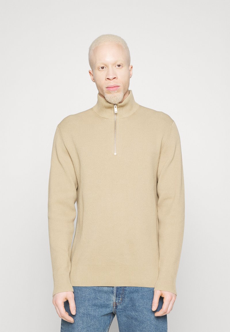 Pullover a coste beige con colletto alto e dettaglio zip, indossato con jeans blu. Il tessuto ha una texture morbida e una vestibilità casual.