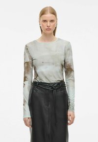 Naine, kellel on sirged blondid juuksed, kannab pikki varrukatega sidrunitehnikas peenvillast pluusi ja kõrge vöökohaga musta nahkse seeliku, seistes valge tausta ees.