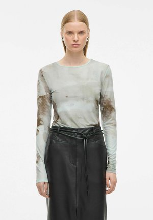 Mujer con cabello rubio lacio viste una blusa de manga larga de gasa con estampado de tie-dye y una falda de cuero negro de tiro alto con cinturón, de pie contra un fondo blanco.