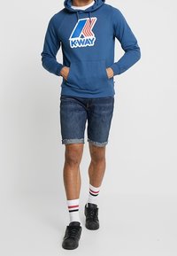 Sininen huppari, jossa on suuri K-Way-logo, yhdistettyinä haalistuneisiin farkkushortseihin, mustiin lenkkareihin ja valkoisiin sukkiin, joissa on punaiset raidat.