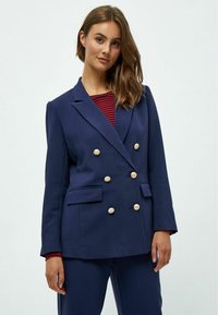 Námořnický dvouřadový blazer se zlatými knoflíky, dvěma předními kapsami, strukturovanými rameny a hladkým povrchem, nošený přes pruhované tričko.