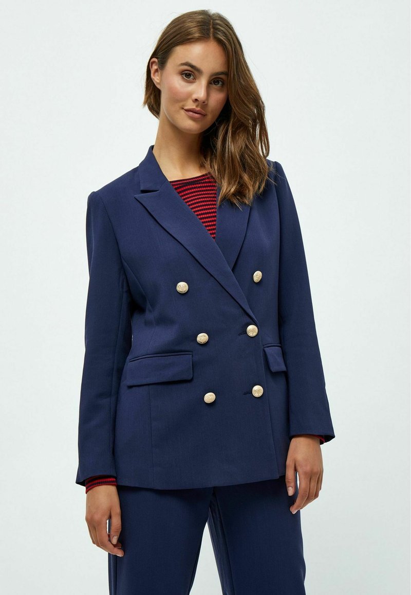 Námořnický dvouřadový blazer se zlatými knoflíky, dvěma předními kapsami, strukturovanými rameny a hladkým povrchem, nošený přes pruhované tričko.