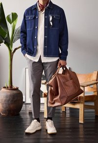 Mörk jeansjacka med två framfickor, vit skjorta med polka-prickar, rutiga byxor, brun läderduffelväska och vita sneakers.