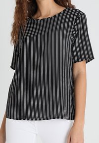 Haut noir à manches courtes avec rayures verticales blanches, encolure ronde et coupe ample. Confectionné en tissu léger ; texture lisse.
