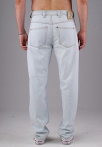 Mann in hellblauen Jeans und weißen Converse-Sneakern, der mit sichtbarem Rücken vor einem einfarbigen grauen Hintergrund steht.