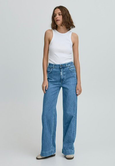 Biely rebrovaný tielko v kombinácii so širokými modrými džínsami s vysokým pásom a miernym opotrebením na leme.