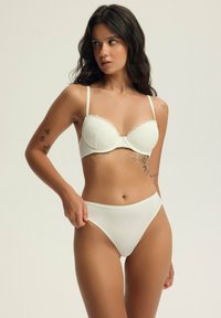Biały koronkowy stanik z falistymi krawędziami i cienkimi ramiączkami, w zestawie z białymi majtkami o wysokim stanem. Gładka tekstura i bezszwowy design.