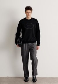 Maglione a maglia nera con logo BOSS testurizzato, abbinato a pantaloni grigi a righe. Il modello tiene in mano una pochette nera, indossando scarpe nere lucide.