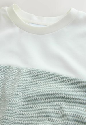 Hvid crewneck-top delvist dækket af et foldet lysegrønt stof med et lille geometrisk mønster.