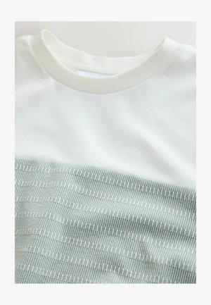 Witte crewneck top, gedeeltelijk bedekt door een opgevouwen lichtgroene stof met een klein geometrisch patroon.